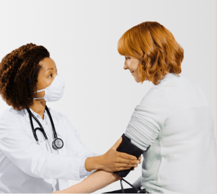 A masked healthcare provider with curly brown hair and wearing a white lab coat gently takes the blood pressure while glancing up at a calm patient who is smiling back.
